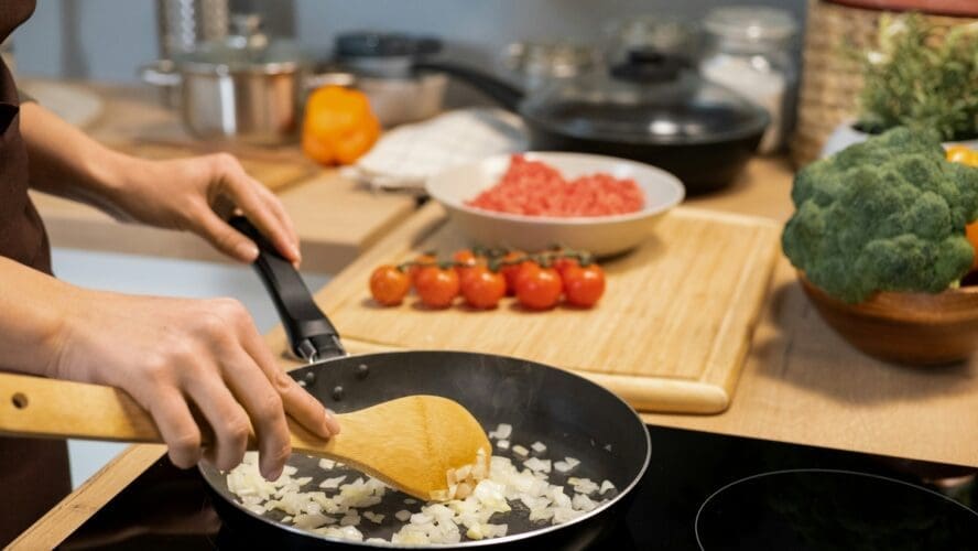 Image of someone cooking onions with a traditional nonstick pan on a glass stovetop, with tomatoes on a cutting board next to them.