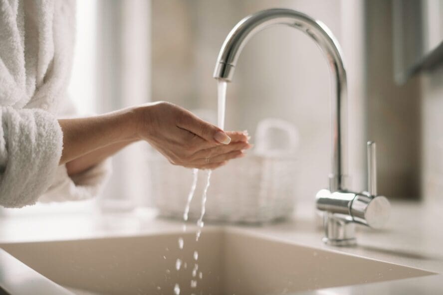 Image of a woman in a bathrobe cupping her hands under running water coming from a sink faucet.