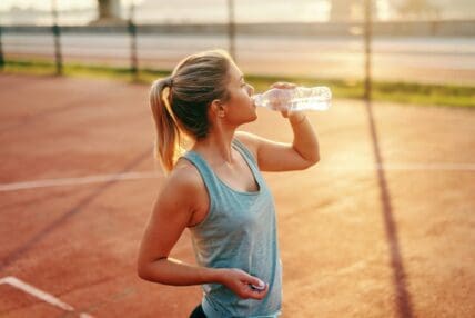 Image of a young blonde woman on a tennis court drinking from a plastic water bottle that likely contains microplastics.