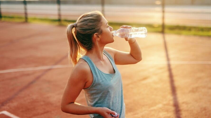 Image of a young blonde woman on a tennis court drinking from a plastic water bottle that likely contains microplastics.