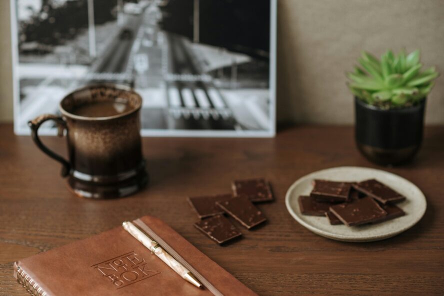Image of several squares of dark chocolate on a plate and on a table set alongside a leather notebook.