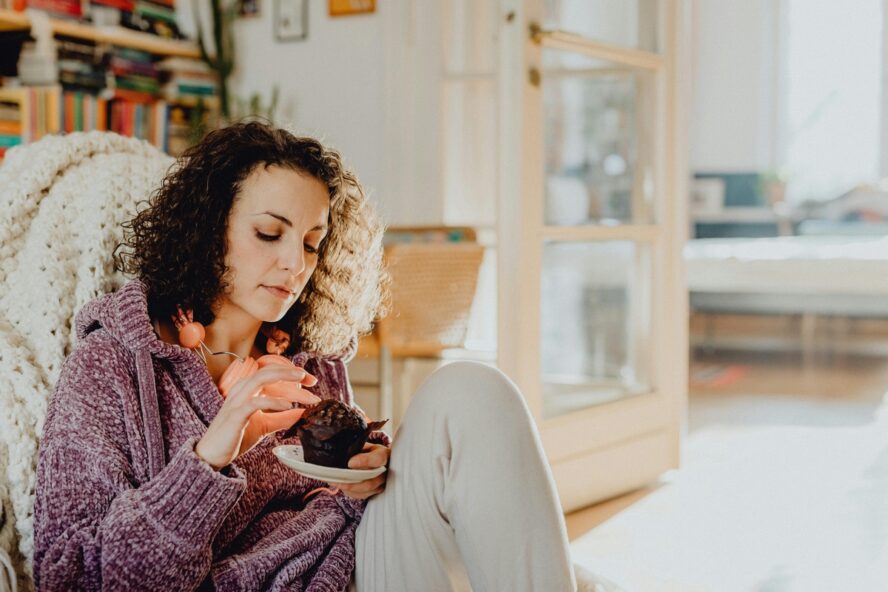 Image of a woman in a cozy sweater sitting against a sofa and picking at a chocolate muffin.