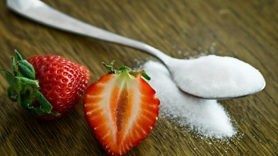 Image of a strawberry sliced in half next to a spoon overflowing with an artificial sweetener like aspartame.