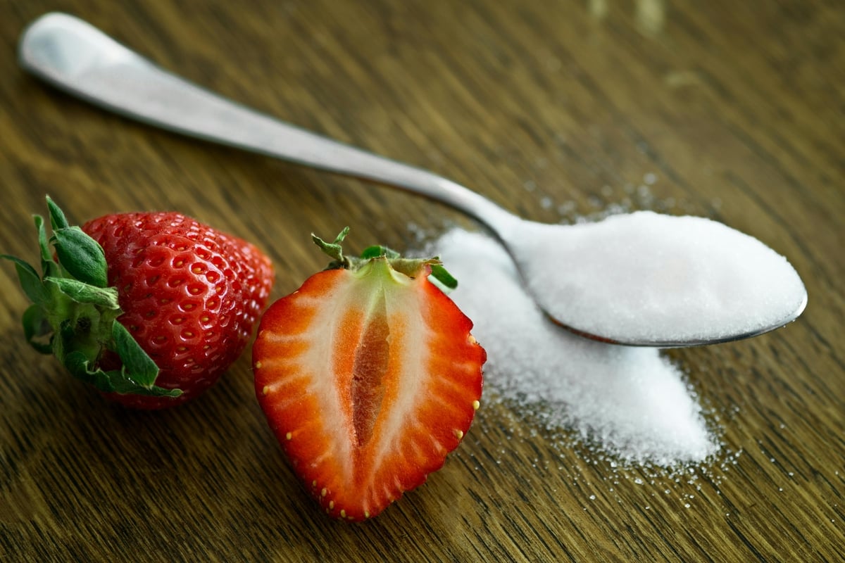Image of a strawberry sliced in half next to a spoon overflowing with an artificial sweetener like aspartame.