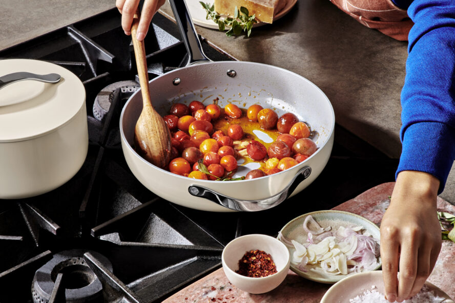 Image of cherry tomatoes in a Caraway saucepan being stirred with a wooden spoon on a stovetop.