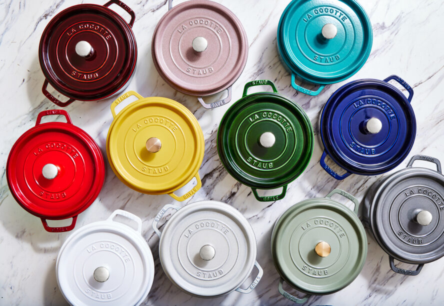 Overhead image of 11 Staub Round Cocottes in a rainbow of colors, from red and yellow to lilac and mint.