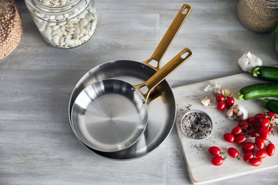 Image of two GreenPan Reserve Stainless Steel fry pans nested inside of each other, next to a cutting board with tomatoes, garlic, and zucchini.