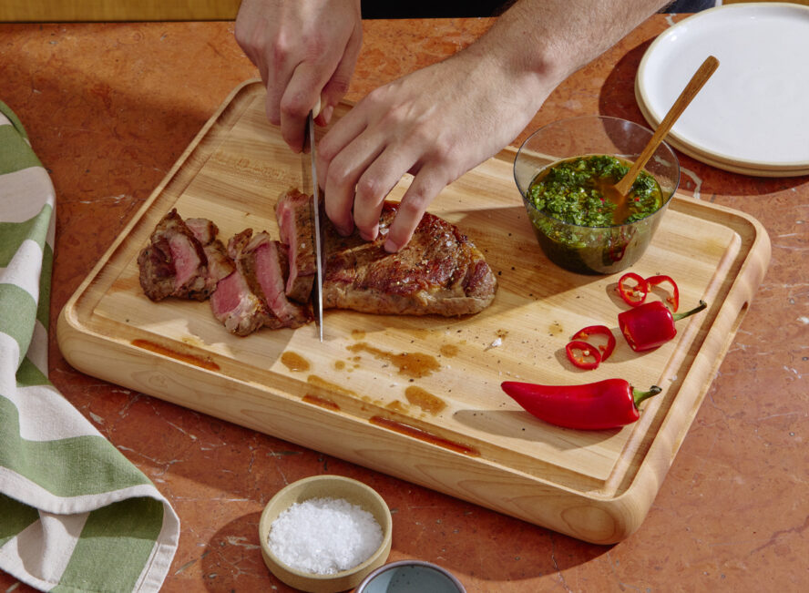 Image of the Caraway Dual-Sided Butcher Block, with a pair of hands slicing a filet steak.