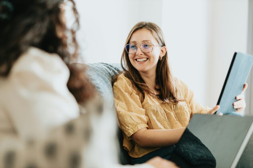Image of a young woman with glasses smiling at another woman in conversation sitting on a couch.