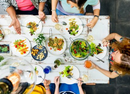 A white dining table shown from overhead with multiple women sitting around it and eating from plentiful bowls of salads and other Mediterranean diet foods.