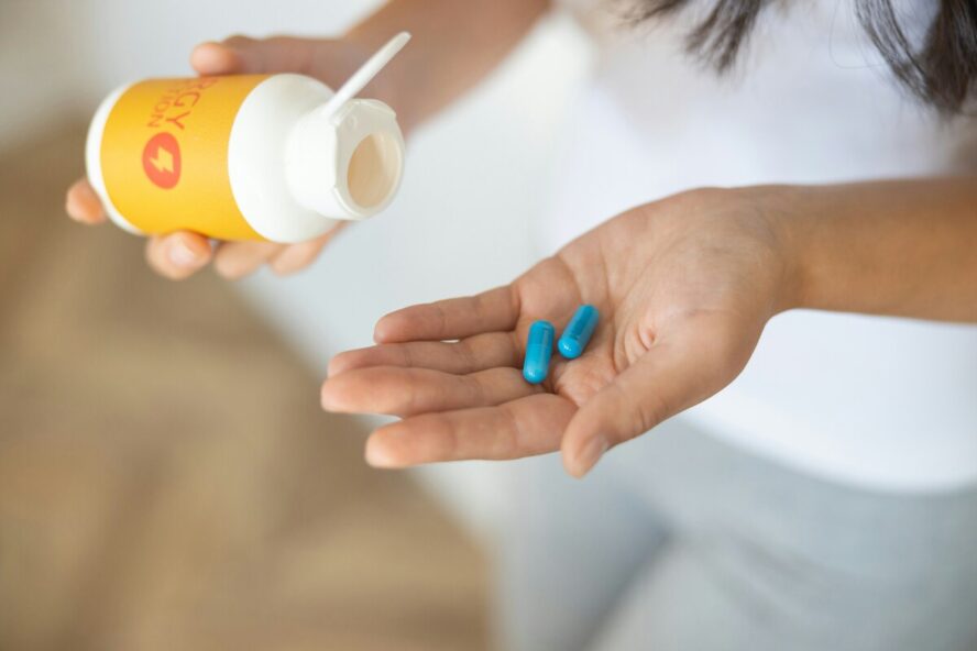 A pair of woman's hand, one hand is holding a bottle of supplements and the other is face up with two blue pills in her palm.