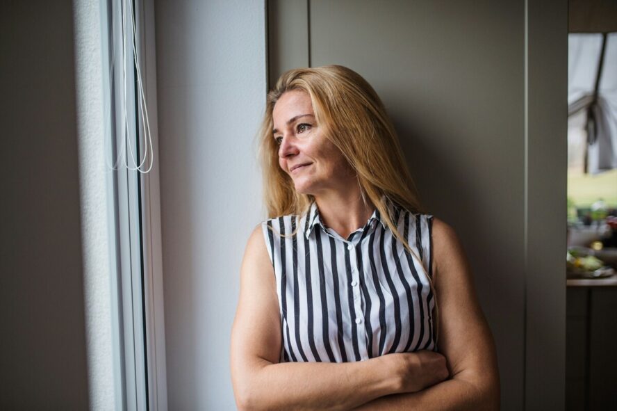 A blonde middle-aged woman standing with her arms crossed, looking out a window with a small smile on her face.