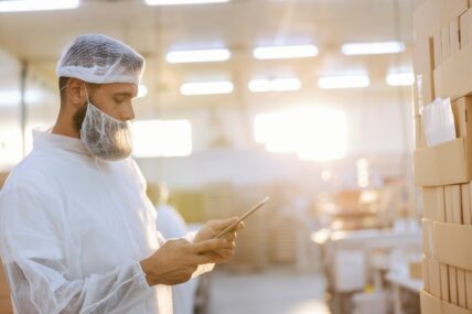 Image of a food safety inspector wearing a hairnet and mask over his beard, holding a tablet in a food production facility.