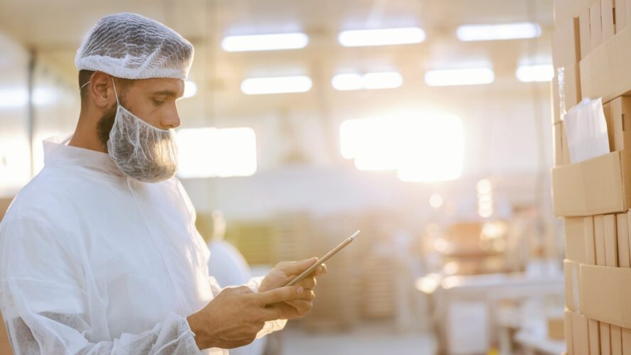 Image of a food safety inspector wearing a hairnet and mask over his beard, holding a tablet in a food production facility.