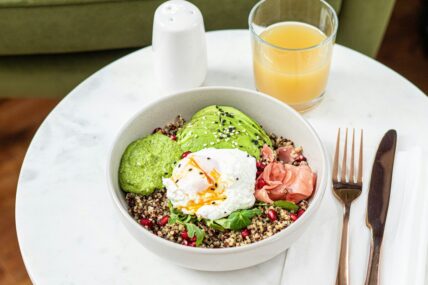 Image of a healthy breakfast bowl full of quinoa, eggs, avocado, pomegranate, and some smoked salmon, alongside gold utensils and a glass of orange juice.