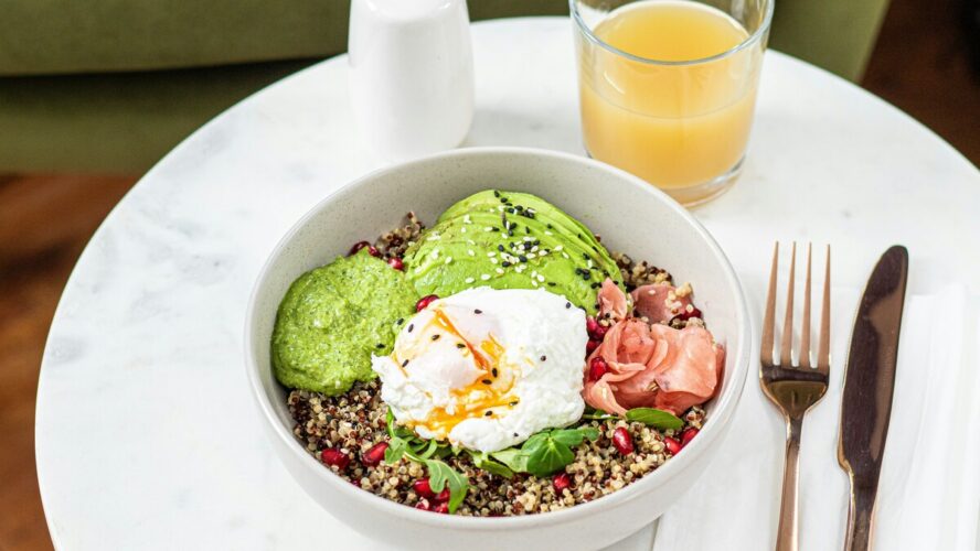 Image of a healthy breakfast bowl full of quinoa, eggs, avocado, pomegranate, and some smoked salmon, alongside gold utensils and a glass of orange juice.
