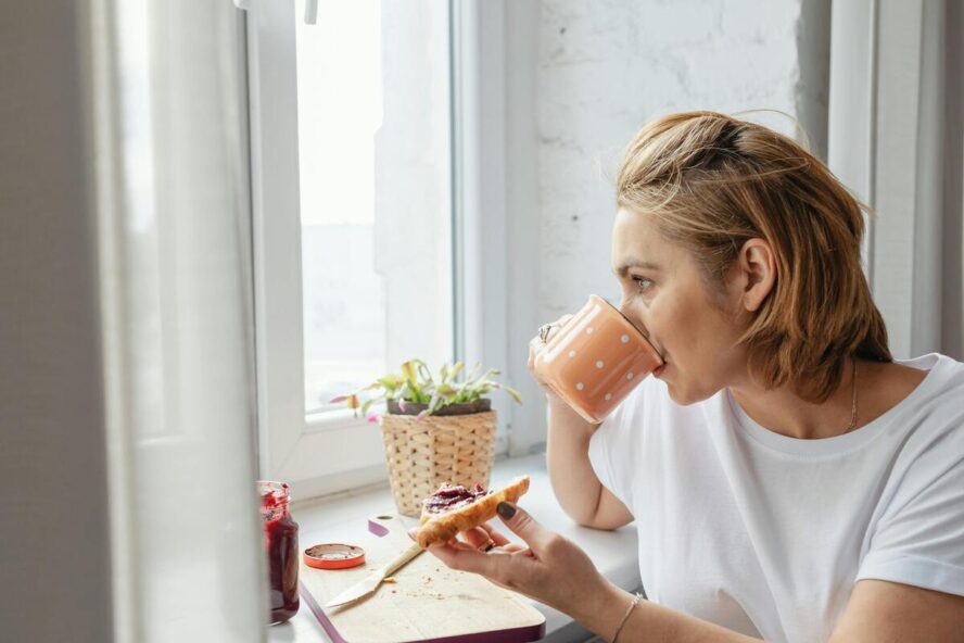 Image of a woman sipping from a mug of coffee and holding a piece of toast in her hand as she gazes out the window.