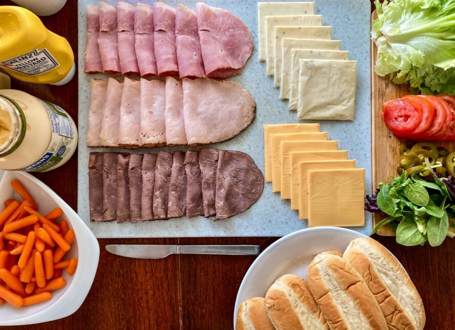 Image of a spread of deli meat cold cuts, various sliced cheeses, and other sandwich ingredients laid out on a board.