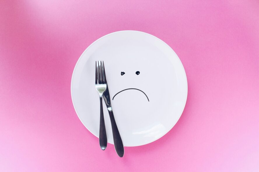 Image of an empty plate of food with a sad face drawn onto it, with a pink background.