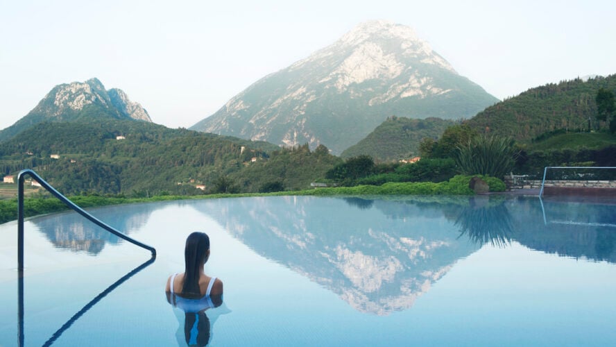 Image of a woman wading into an infinity pool overlooking the mountains of the Eastern Alps.
