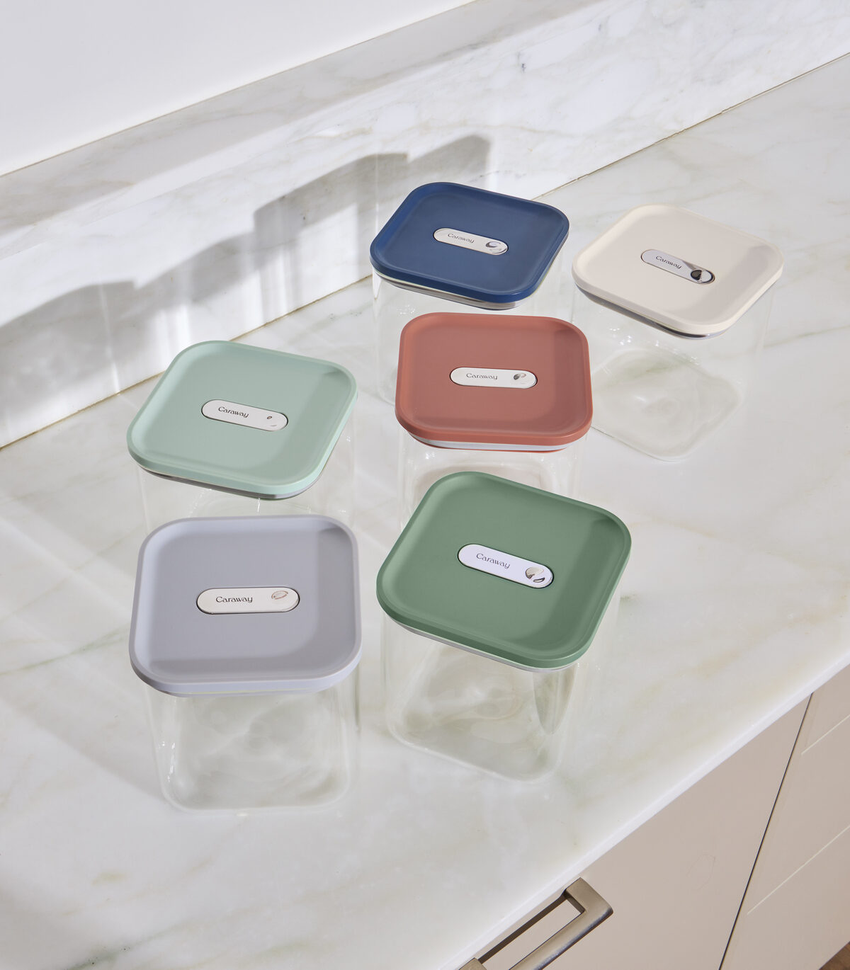 Image of the Caraway Glass Airtight Container set with various different color lids displayed on a white marble countertop.