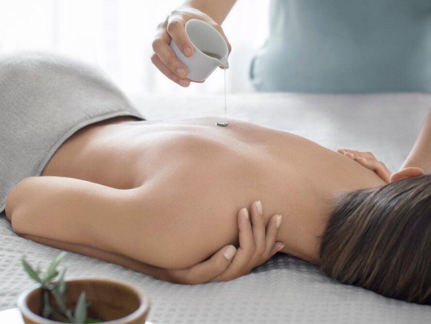 Image of a woman lying face-down on a massage table, with a masseuse pouring oil over her back.
