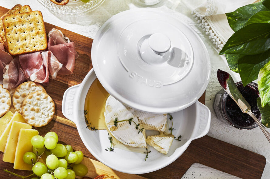 Image of the Staub Round Covered Brie Baker in pure white, filled with a wheel of brie and surrounded by crackers and grapes.