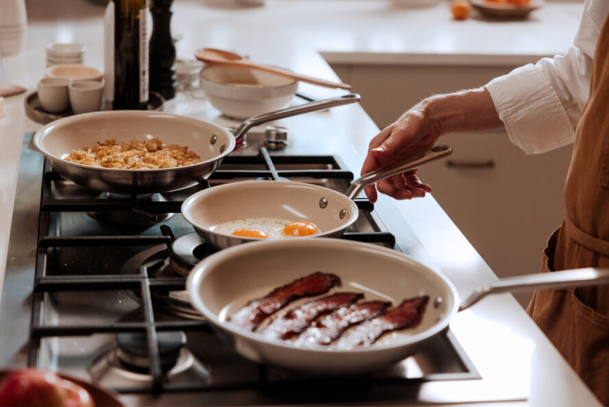 Image of a pair of hands using three Made In CeramiClad frying pans on a stovetop, filled with breakfast items like eggs and bacon.