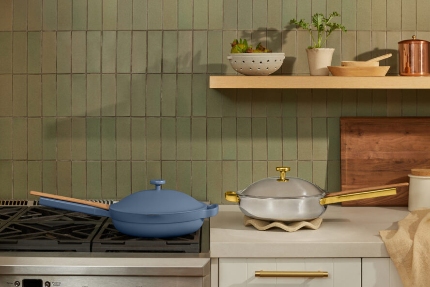 Image of two Our Place pans, one in their classic ceramic nonstick and one Titanium Always Pan Pro in a kitchen with a green tile backsplash.