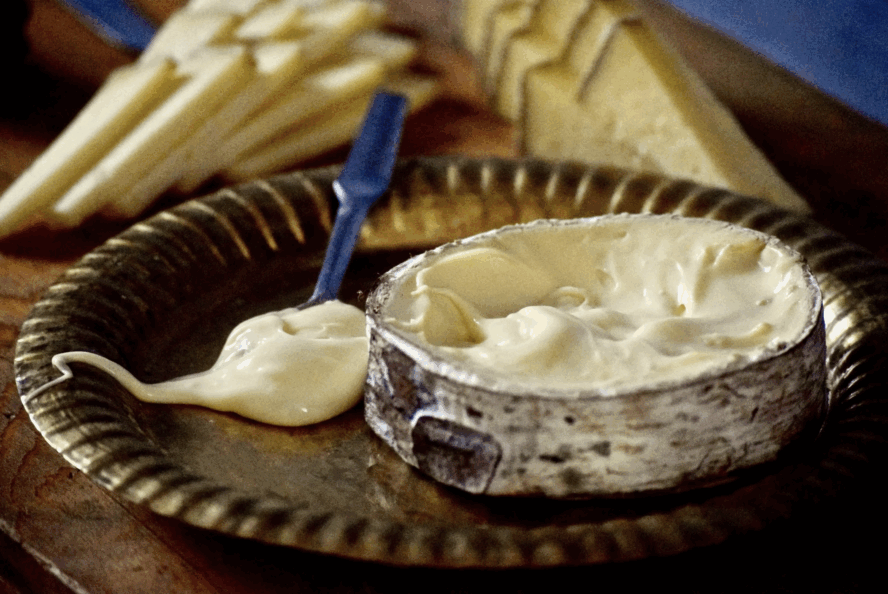 Image of a round of Harbison cheese, with a blue spoon being used to spread the cheese out of its rind.