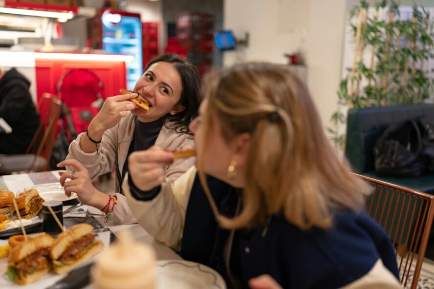 Image of two young women sitting in a fast food restaurant chatting while eating burgers and pizza.