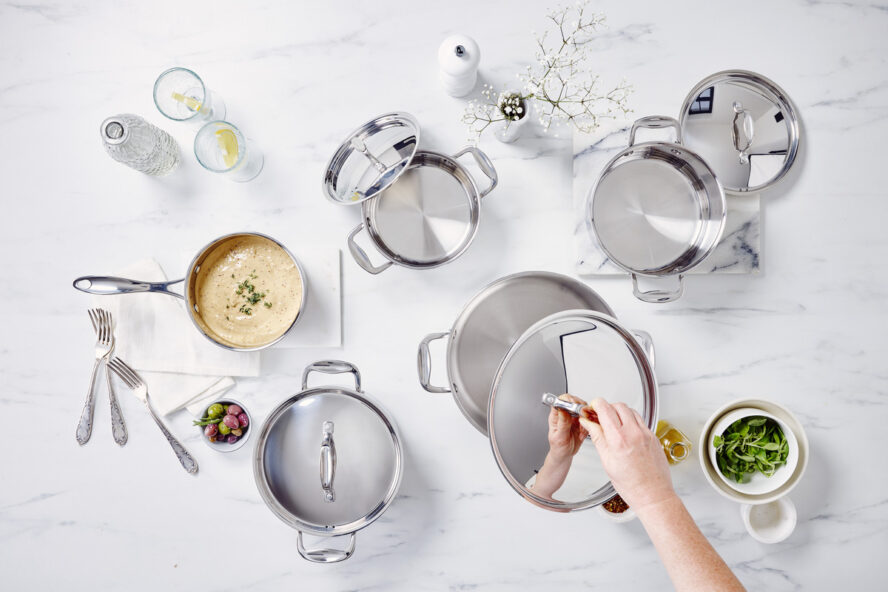 Image of a full set of Alva Stainless Steel cookware shot from above, with a hand reaching in to lift a lid off a pot.