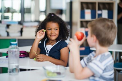 Image of two schoolchildren sitting at schooldesks and comparing snacks — an apple and grapes.