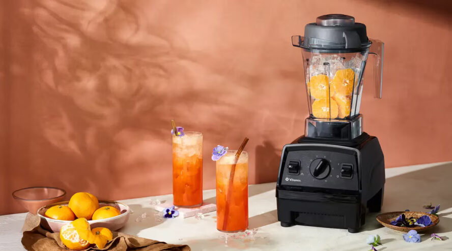 Image of a Vitamix blender filled with citrus and ice, with two glasses of blended juice alongside it.
