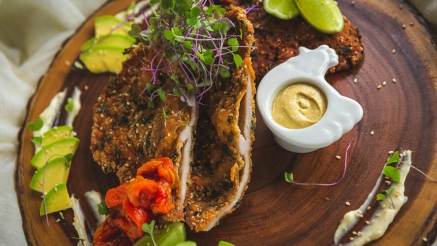 Image of a wooden board with a fried chicken cutlet, mustard, and avocado around it.