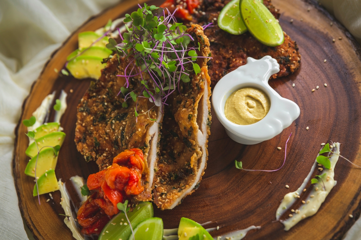 Image of a wooden board with a fried chicken cutlet, mustard, and avocado around it.