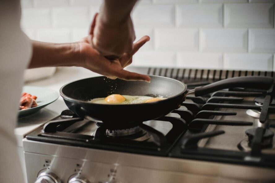 Image of a nonstick pan on the stovetop frying eggs, with a pair of hands grinding salt over them.
