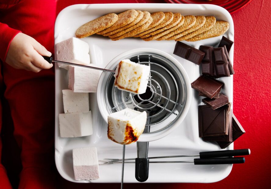 Image of the Williams Sonoma S'mores Maker shown on a red background with a child's hand roasting marshmallows over the flame.