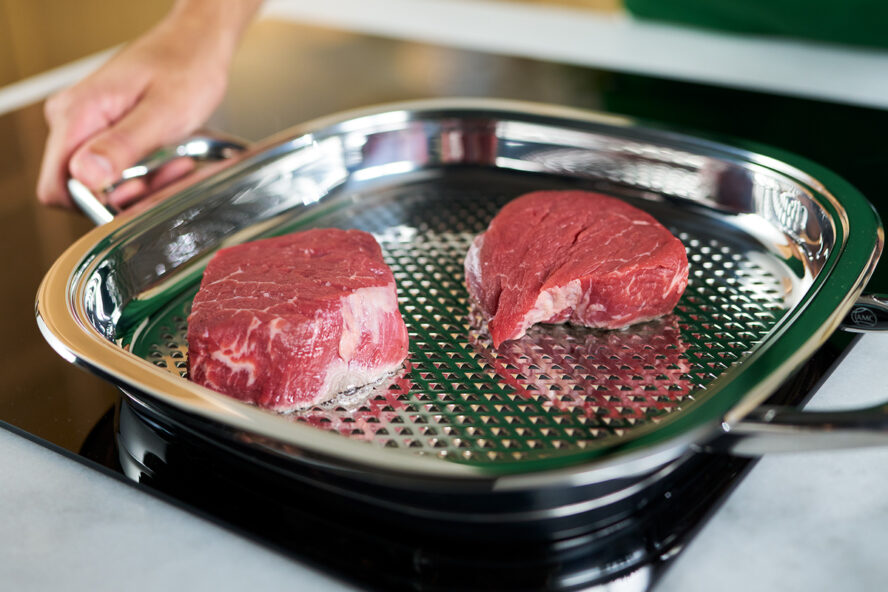 Image of the AbyAMC stovetop grill pan with lid, with a hand holding one handle and searing two steaks.