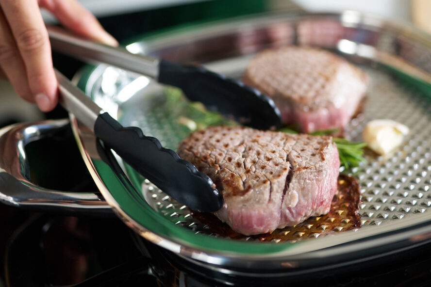 Image of the AbyAMC grill pan with a pair of hands using tongs to flip two fillet steaks.