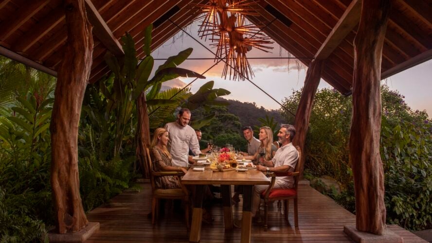 Image of a group sitting at a large dining table overlooking palm trees, with a chef serving them dinner.