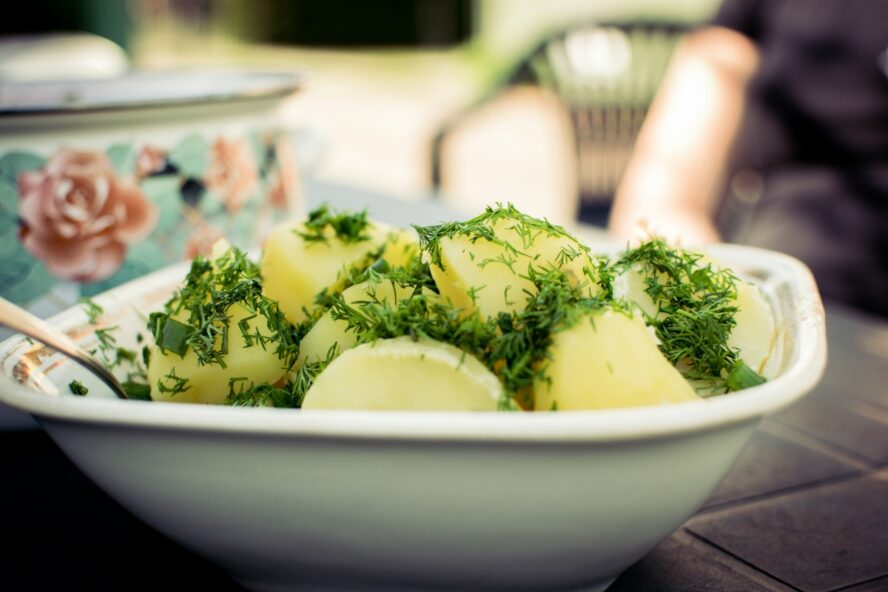 Image of a boiled potato salad with dill in a large white dish on a picnic table.