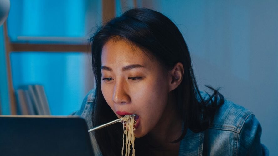 Image of a young woman eating ramen noodles while watching something on a laptop screen.