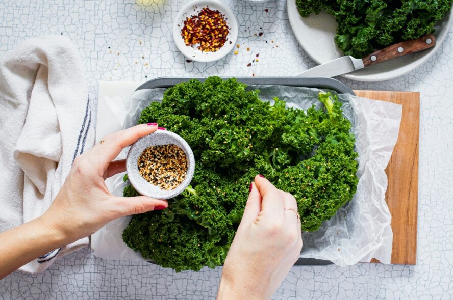 Image of a tray with a pile of kale, with a pair of hands seasoning the high-fiber greens.