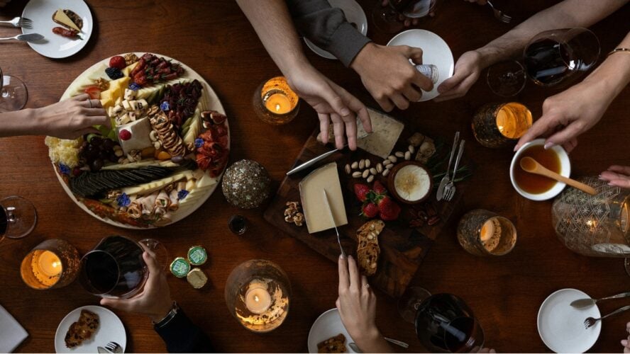 Image of a festive holiday table spread with a cheese board, featuring hands reaching over to help themselves.