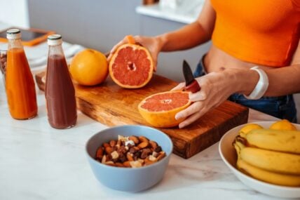 Image of a woman wearing an orange tank top and slicing a grapefruit in half on a cutting board in a brightly lit kitchen.