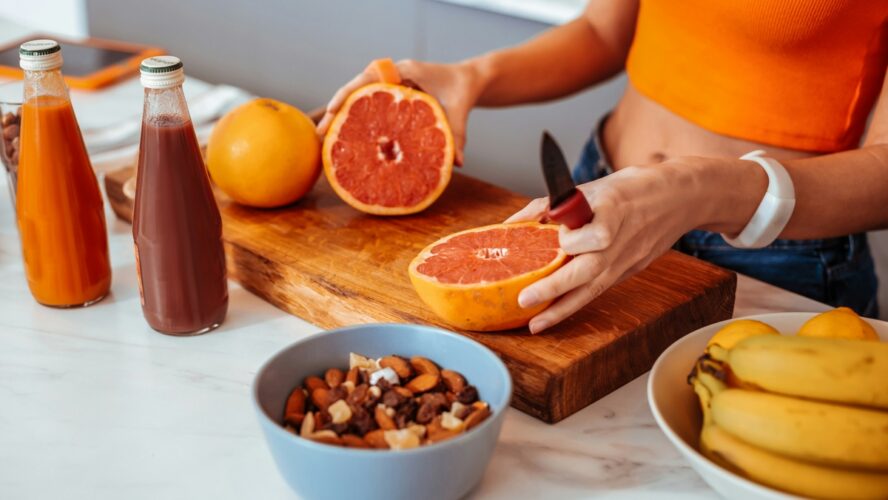Image of a woman wearing an orange tank top and slicing a grapefruit in half on a cutting board in a brightly lit kitchen.