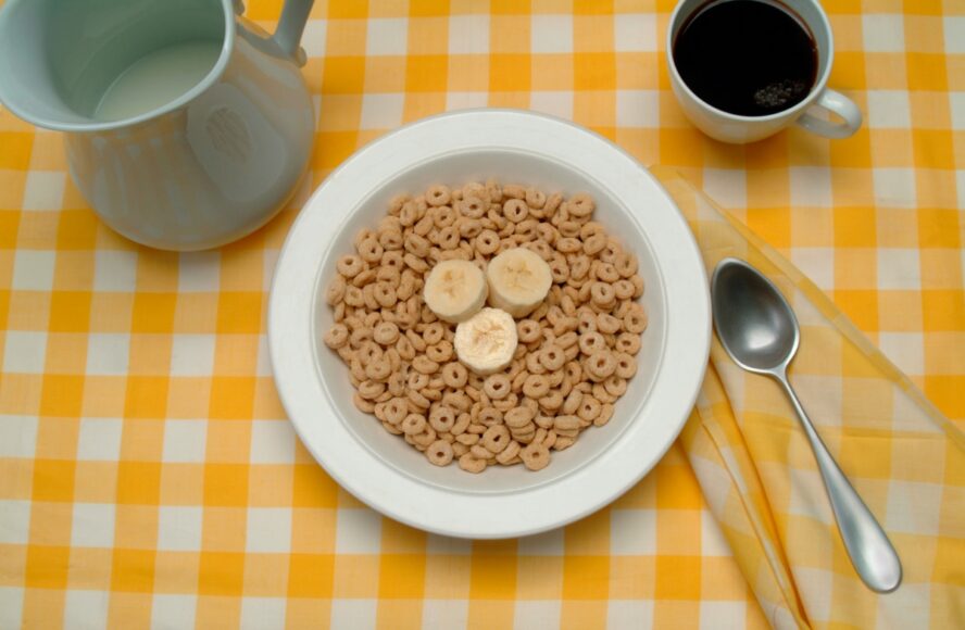 Image of a bowl of Cheerios with some slices of banana on top, shown on a yellow plaid tablecloth.
