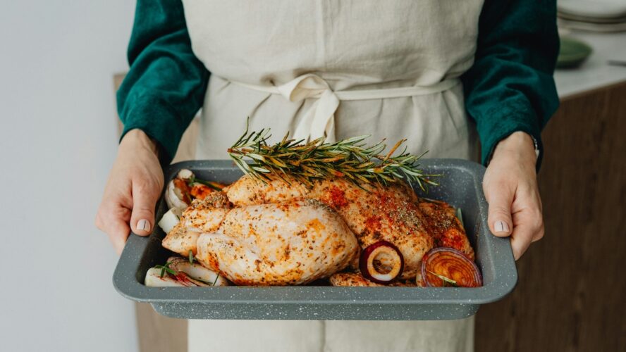 Image of a woman wearing an apron holding an oven tray with a raw chicken, seasoned and topped with rosemary.