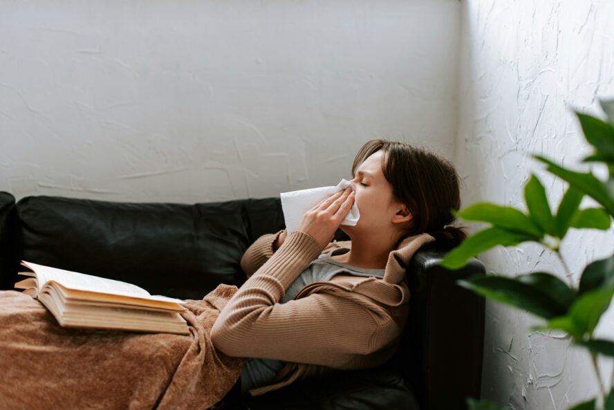 Image of a woman lying down on the couch, blowing her nose with a book in her lap.